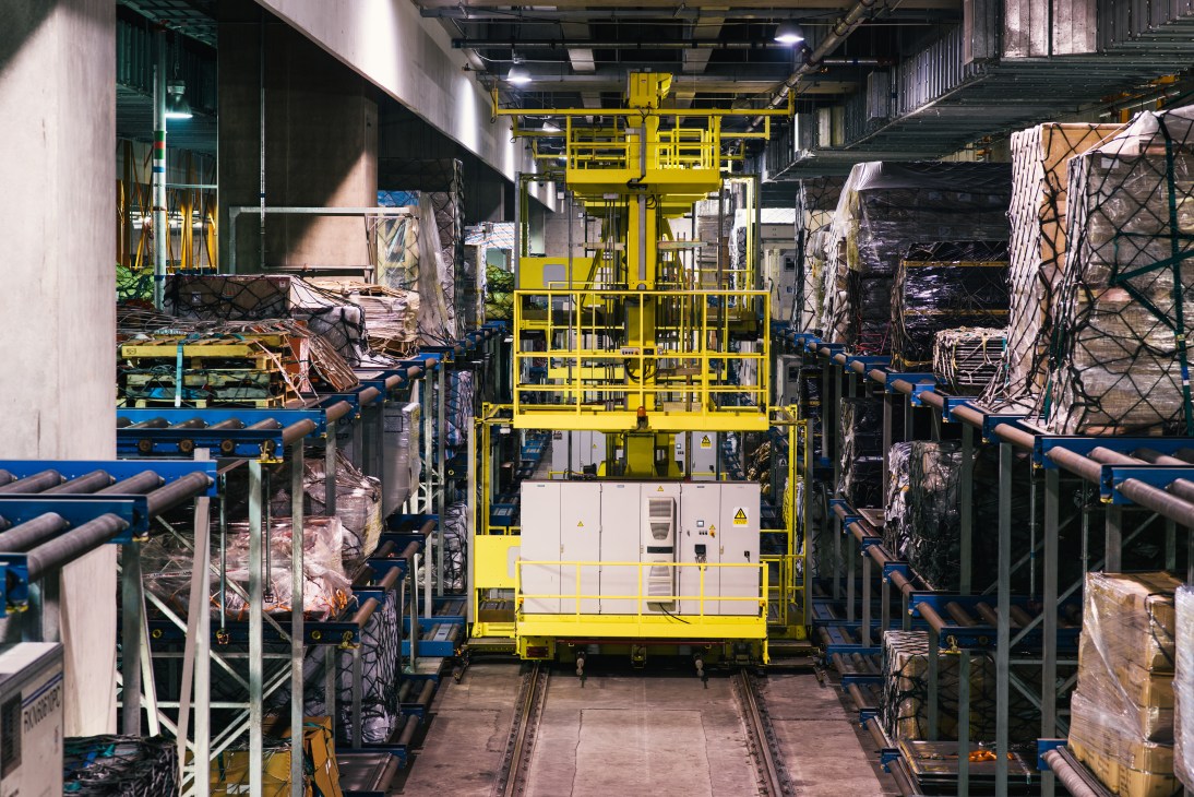 Automated cargo handling system with yellow machinery moving containers on blue conveyor rails in airport terminal.