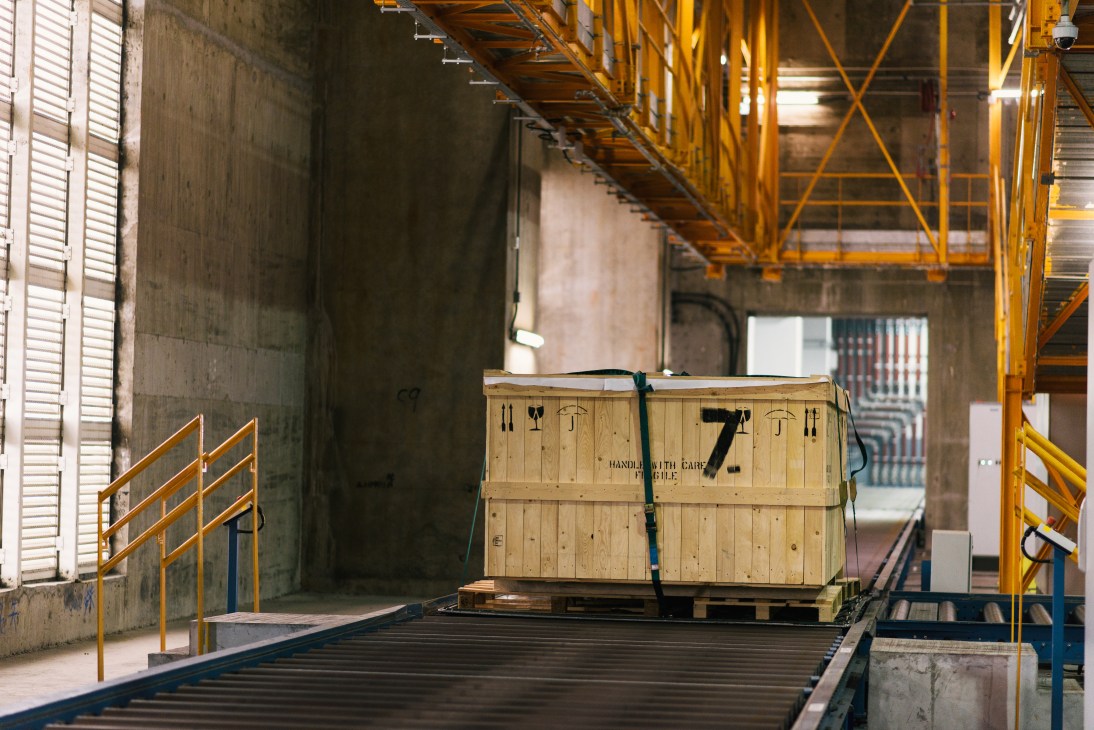 Wooden cargo container on conveyor system beneath yellow industrial machinery in warehouse.