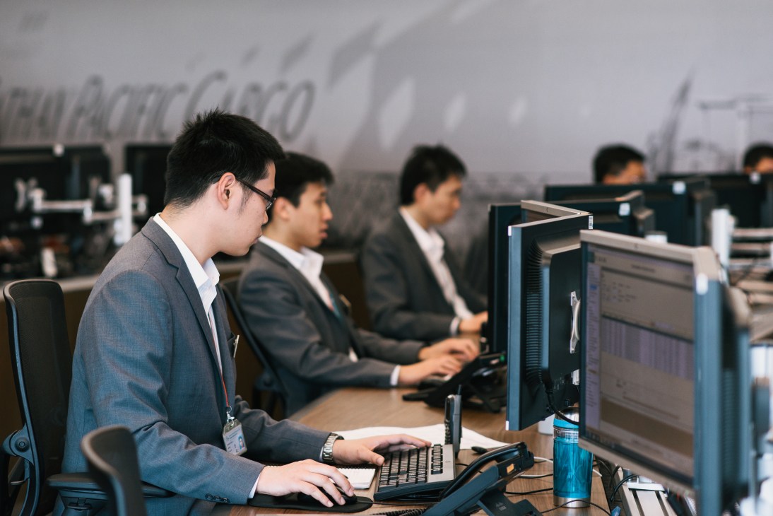 Control room technicians in business attire operating computer systems to manage cargo movement at the airport.