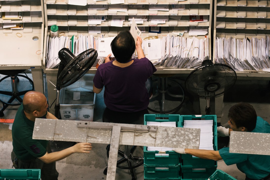 Workers manually checking registered mail at sorting stations with shelves of envelopes and green crates.