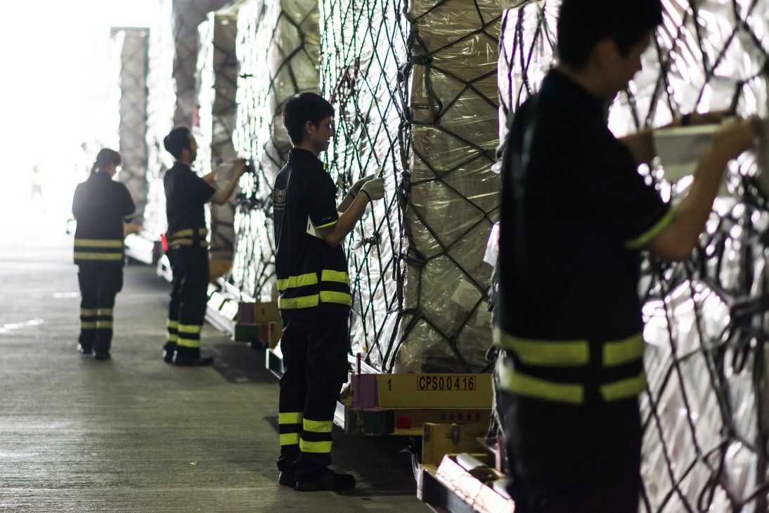 Workers in safety vests securing netted cargo pallets at Hong Kong airport's Quick Transhipment handling area.