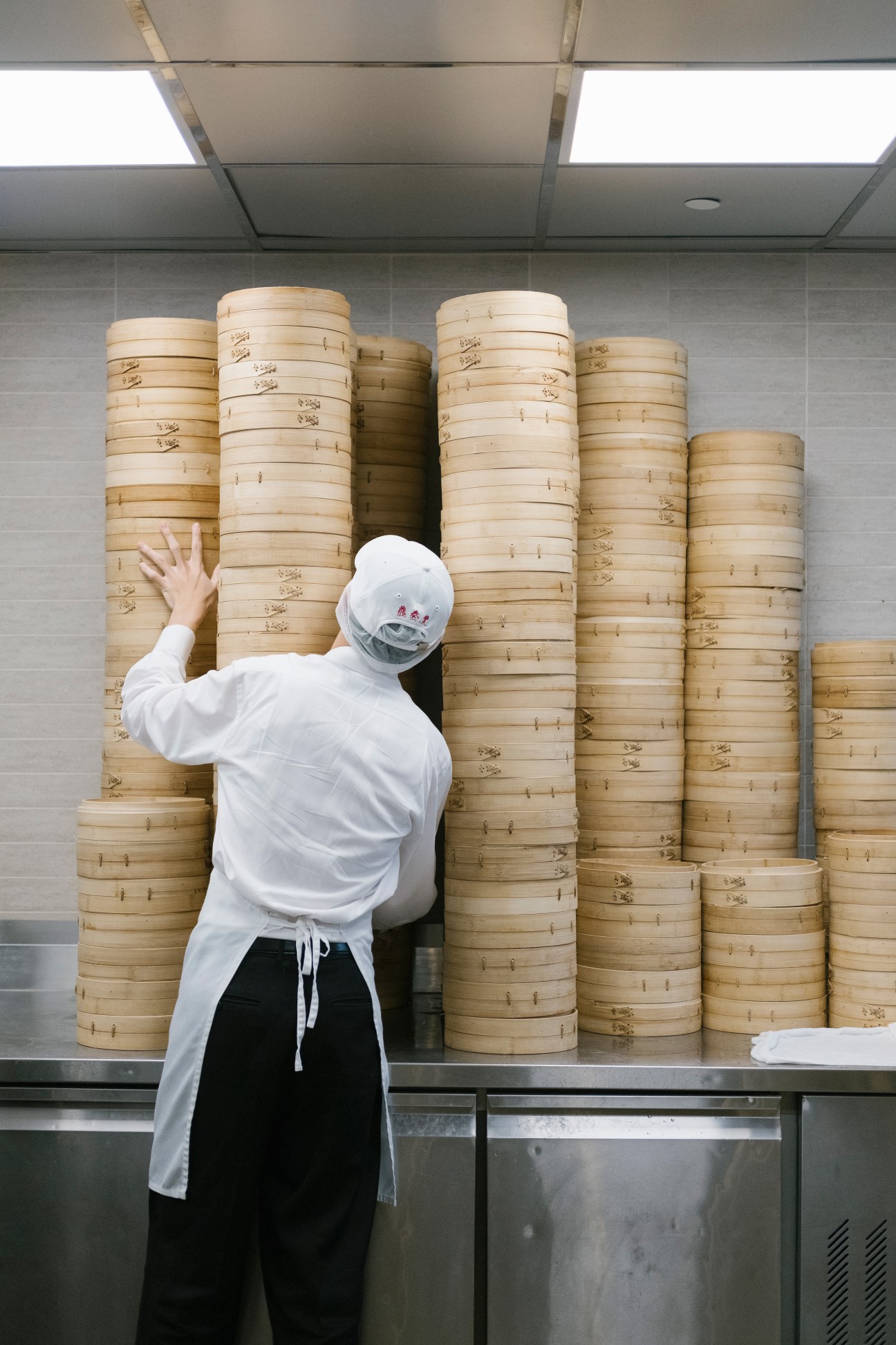 A worker in white chef's coat and mask reaches toward stacked bamboo steamer baskets in a kitchen.