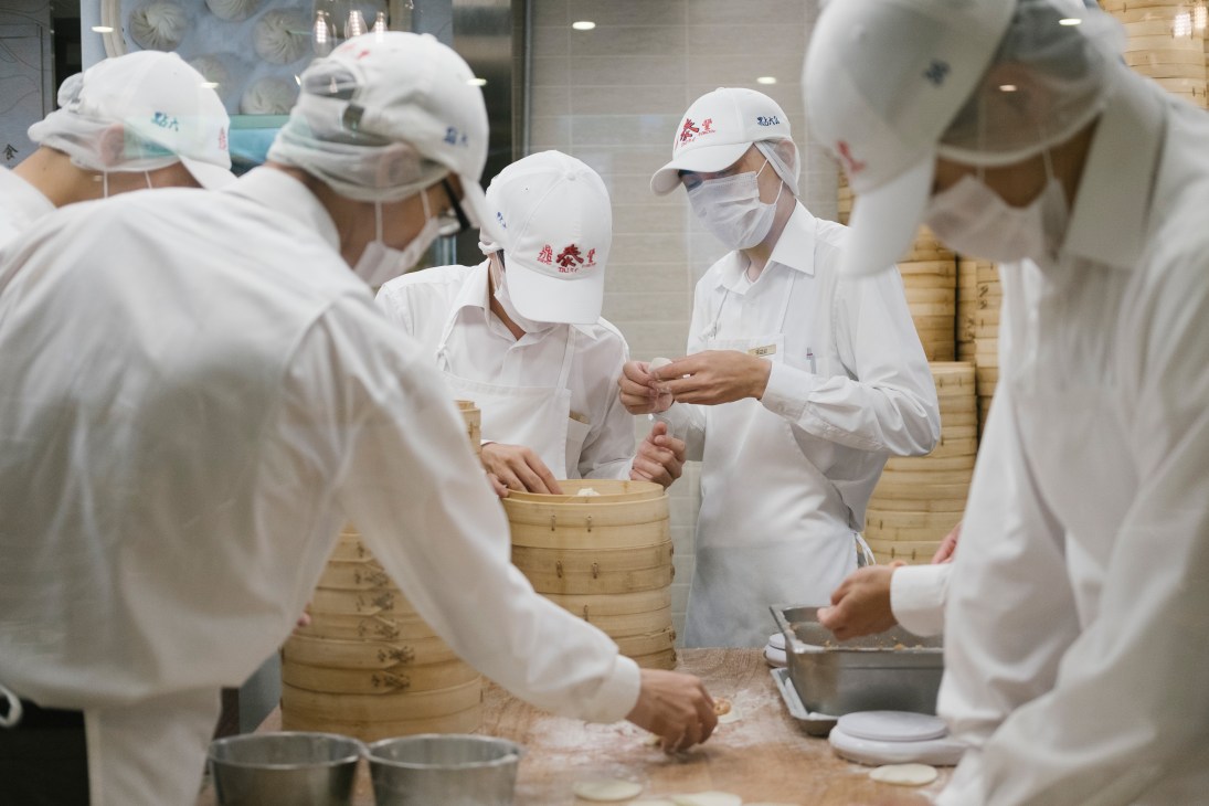 Din Tai Fung staff in white uniforms and caps preparing dumplings in bamboo steamers.
