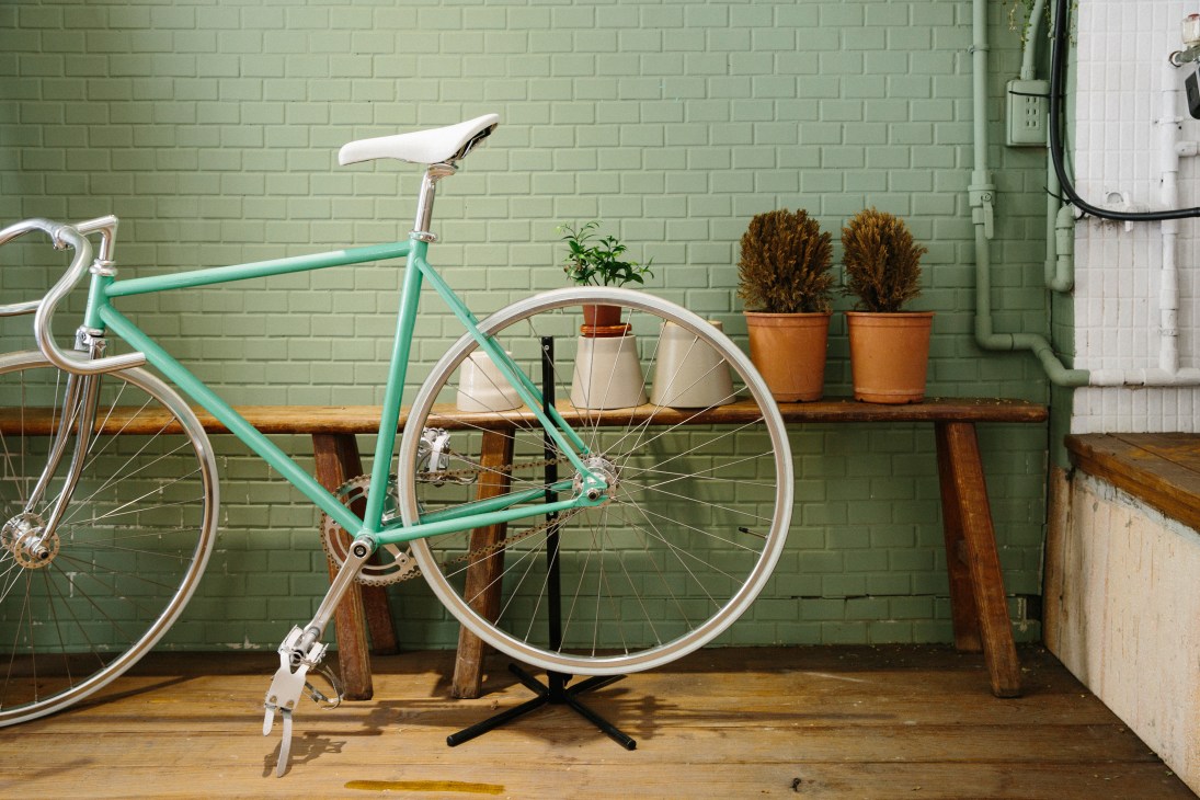 Turquoise bicycle leaning against wooden bench with potted plants against green brick wall.
