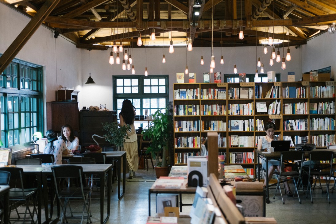 Interior of Yue Yue & Co bookshop with wooden bookshelves, hanging pendant lights, and customers seated at tables.
