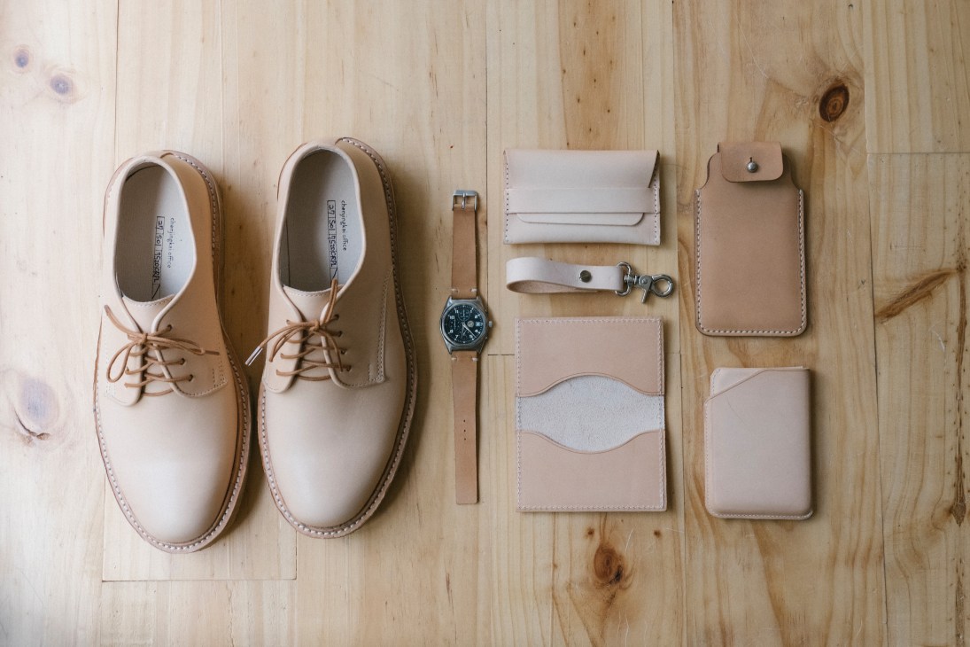 Tan leather shoes and accessories arranged on light wood, including oxfords, watch, belt, and leather goods from Chen Jingka.