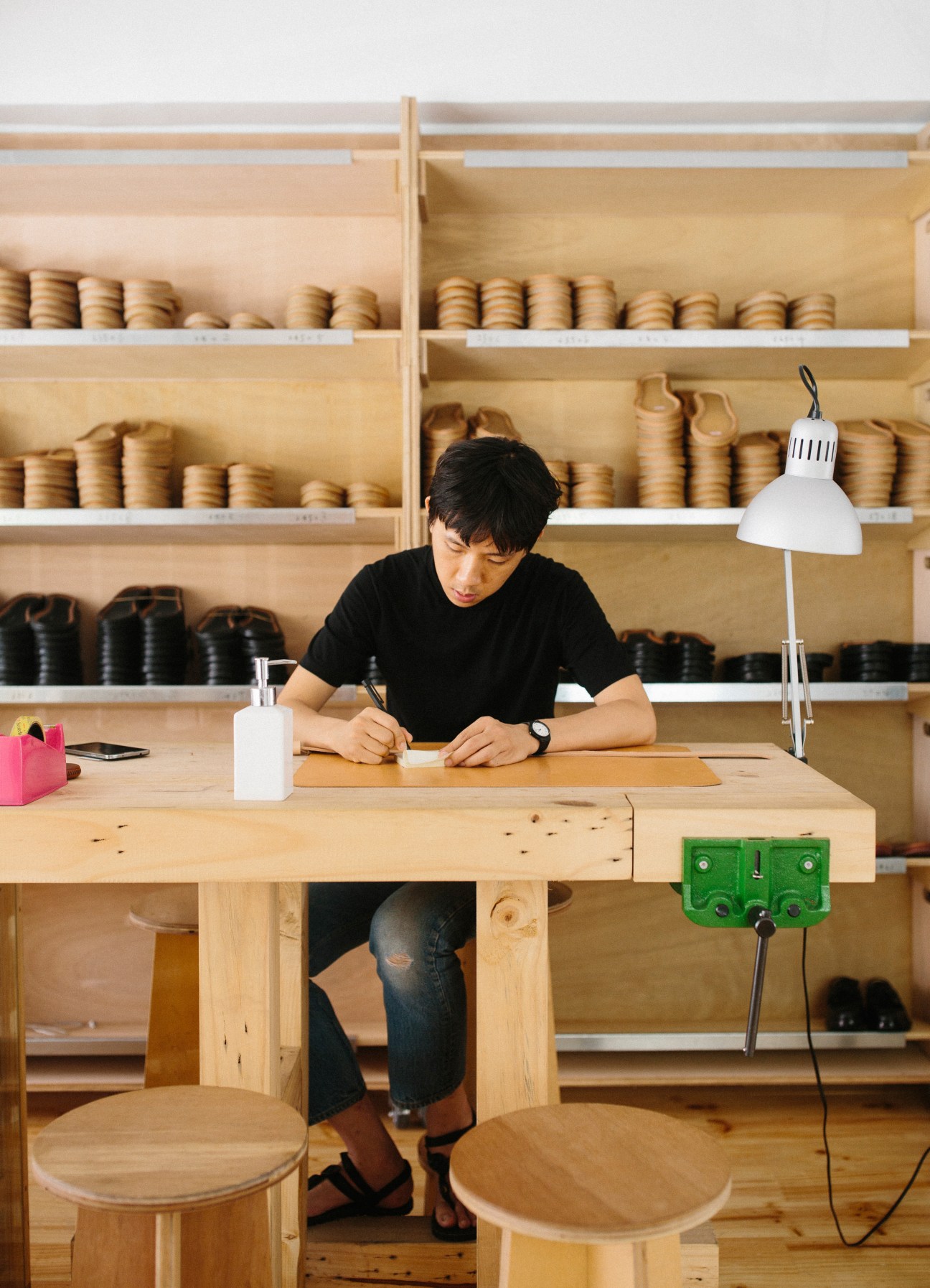 A craftsman at Chen Jingkai footwear shop designs custom shoes at a wooden desk with shelves of shoe components behind him.