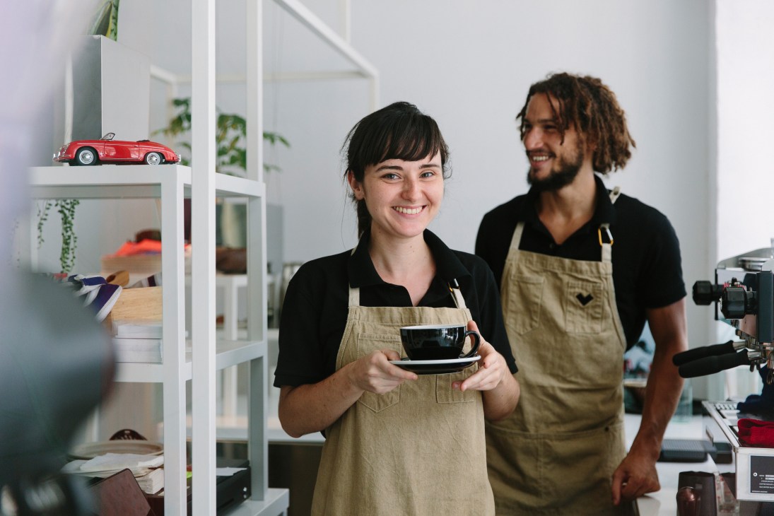 Two staff members in tan aprons smiling in a bright, minimalist cafe interior with shelving and plants.