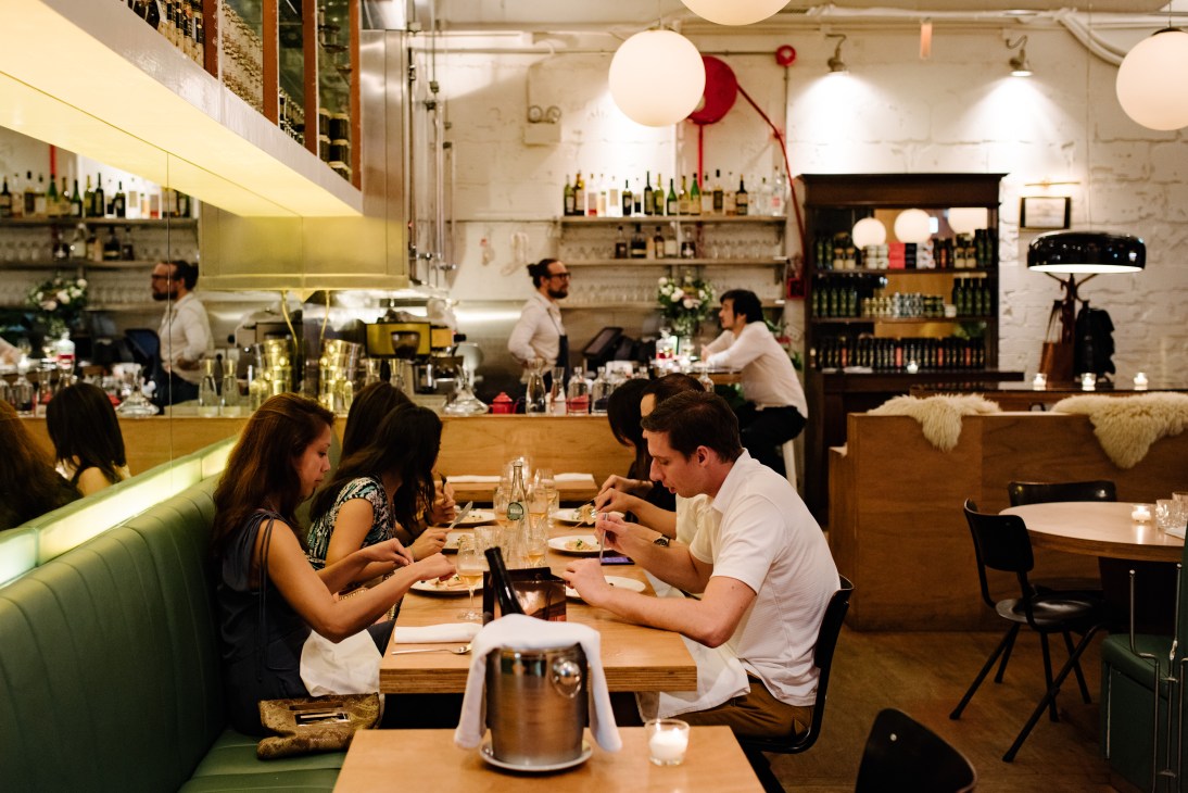 Diners seated at wooden tables in a modern restaurant dining room with green banquettes and white brick walls.