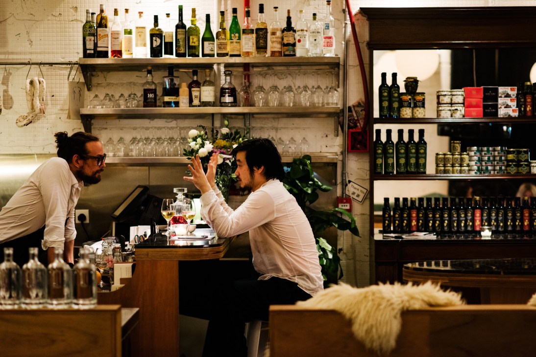 Two men in white shirts chat at a bar with wine bottles and glassware displayed on shelves behind them.