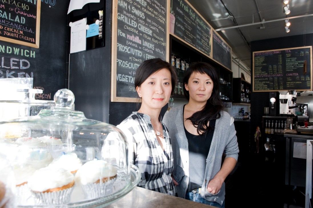 Two women stand behind the counter at Little Branch Cafe, with menu boards and pastries visible.
