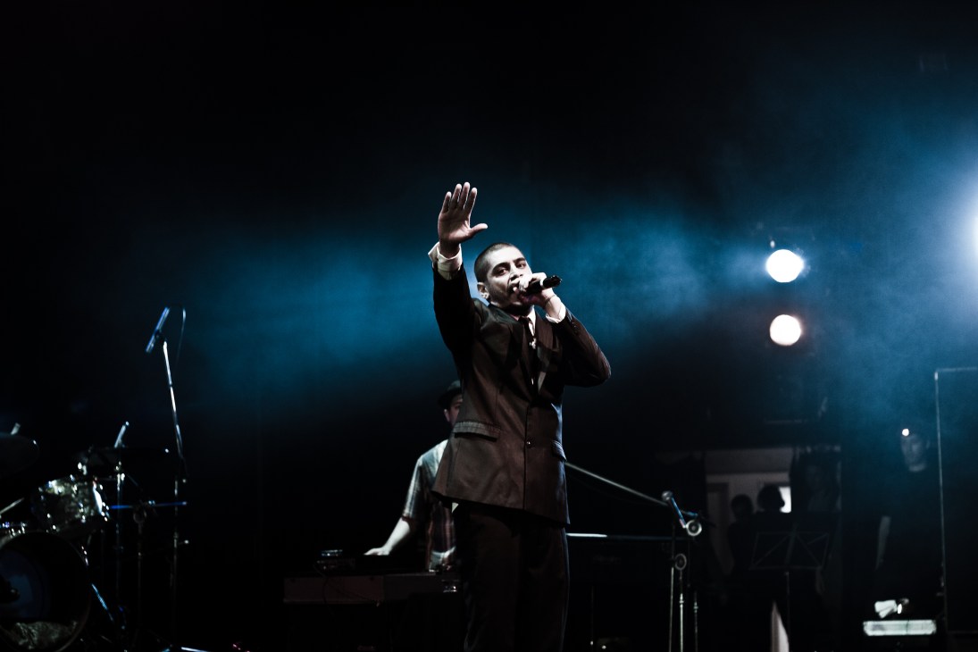 Criolo performing on stage with microphone, raising his hand to the audience under blue stage lighting.