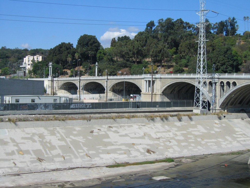 Concrete-lined Los Angeles River with arched bridge, power transmission tower, and hillside vegetation.