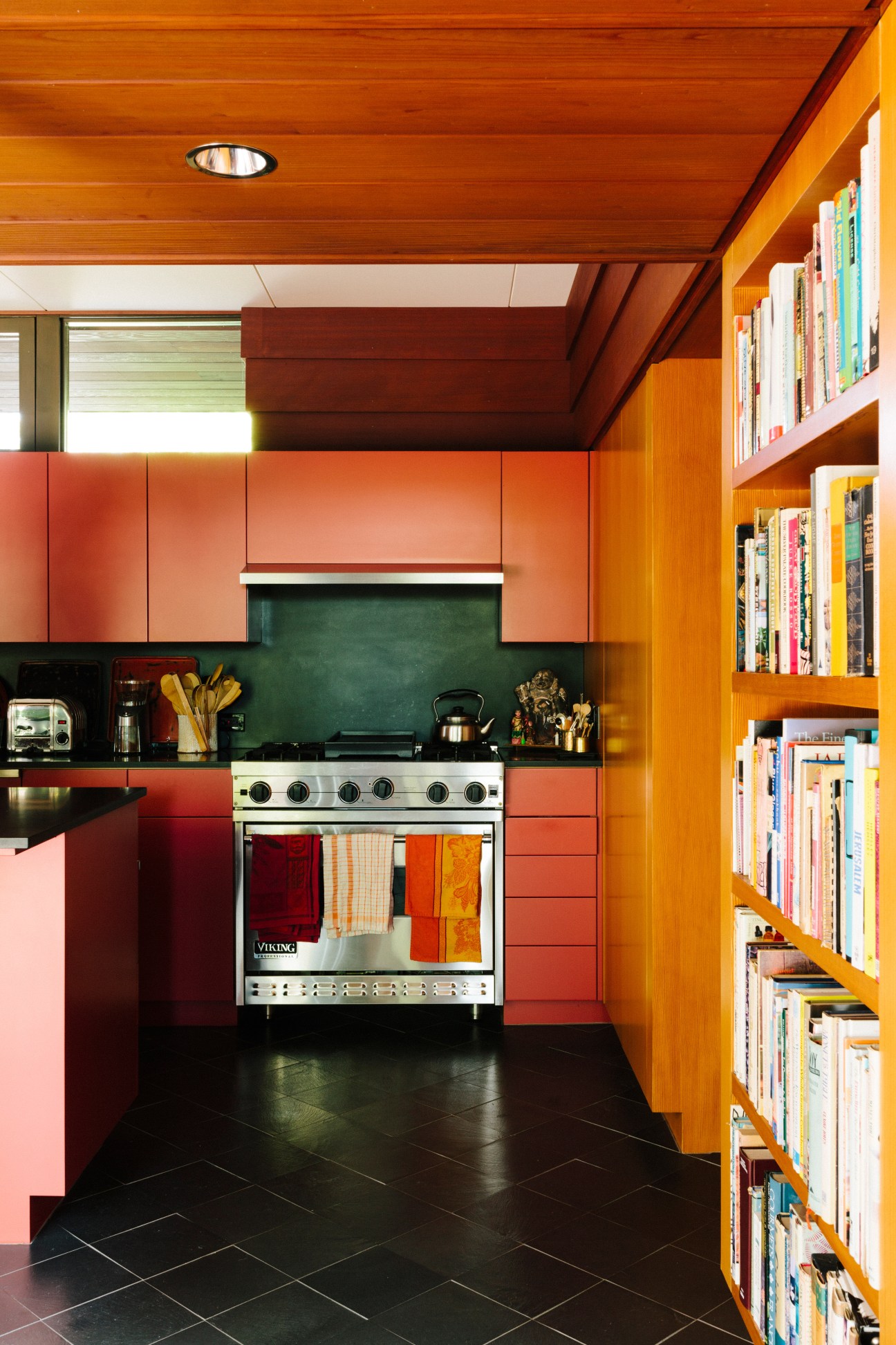 Red kitchen with Viking range, green backsplash, and wood shelving holding cookbooks on the right wall.