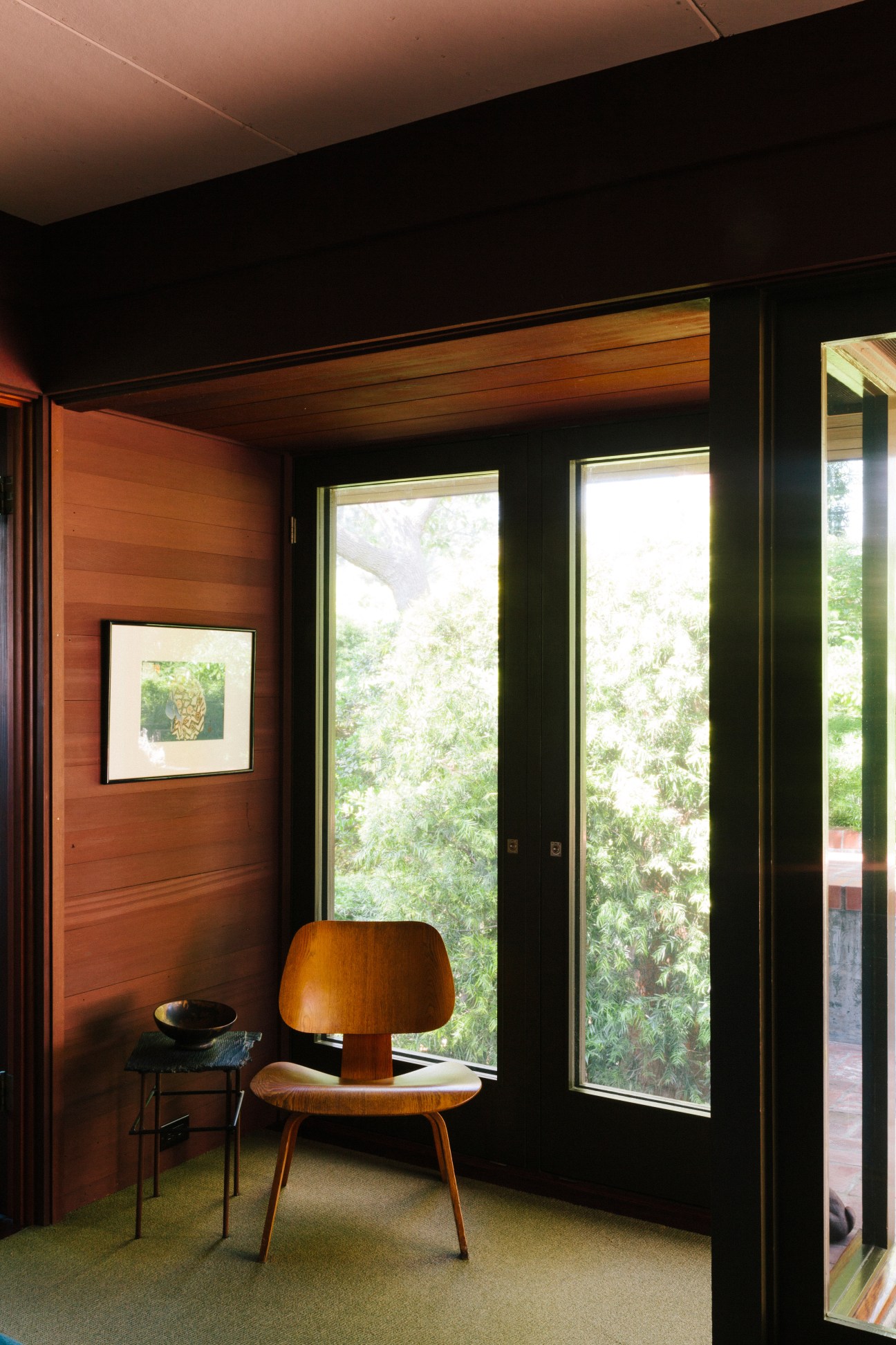Wooden Eames DCW chair positioned by large windows overlooking green foliage in a modernist bedroom.