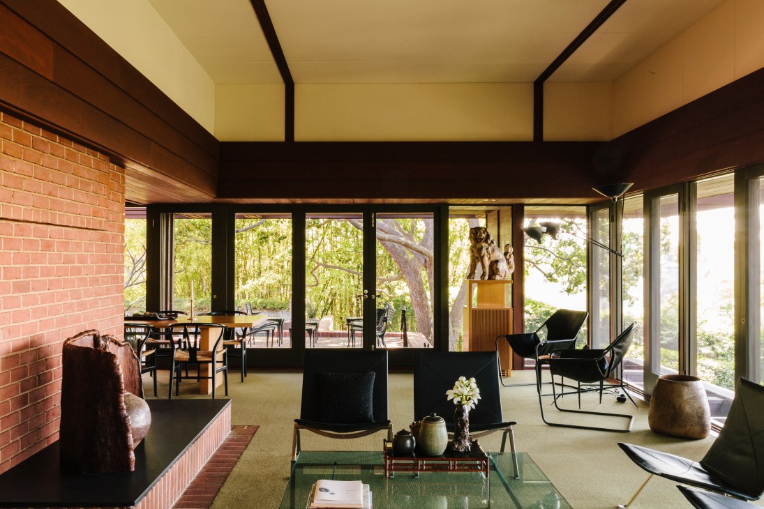 Mid-century modern living room with black chairs, glass table, and expansive views of forest landscape through floor-to-ceiling windows.