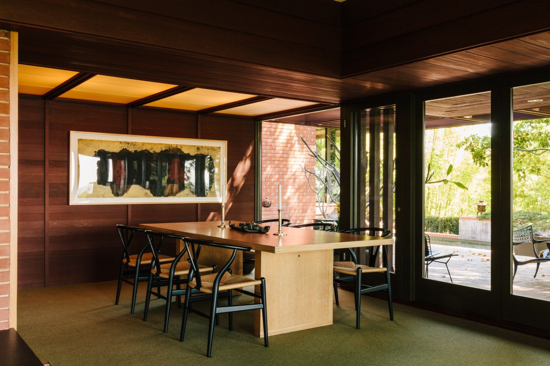 Modern dining area with light wood table, black chairs, and framed abstract artwork on dark wood-paneled wall with warm ceiling lighting.