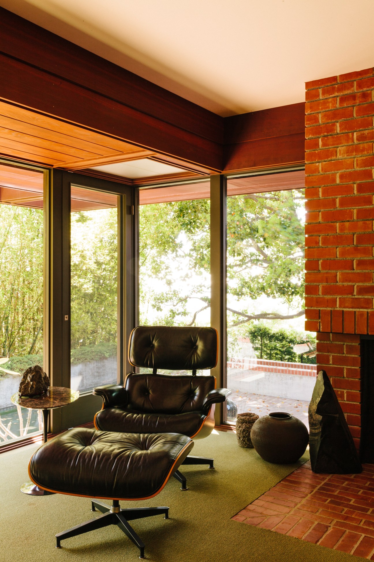 Black leather Eames Lounge Chair with ottoman beside a brick fireplace with large glass doors opening to a garden.