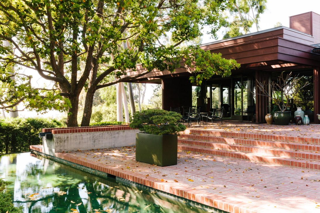 Modern brick patio with a lap pool, shaded by large mature trees, adjacent to a dark-clad residential building.