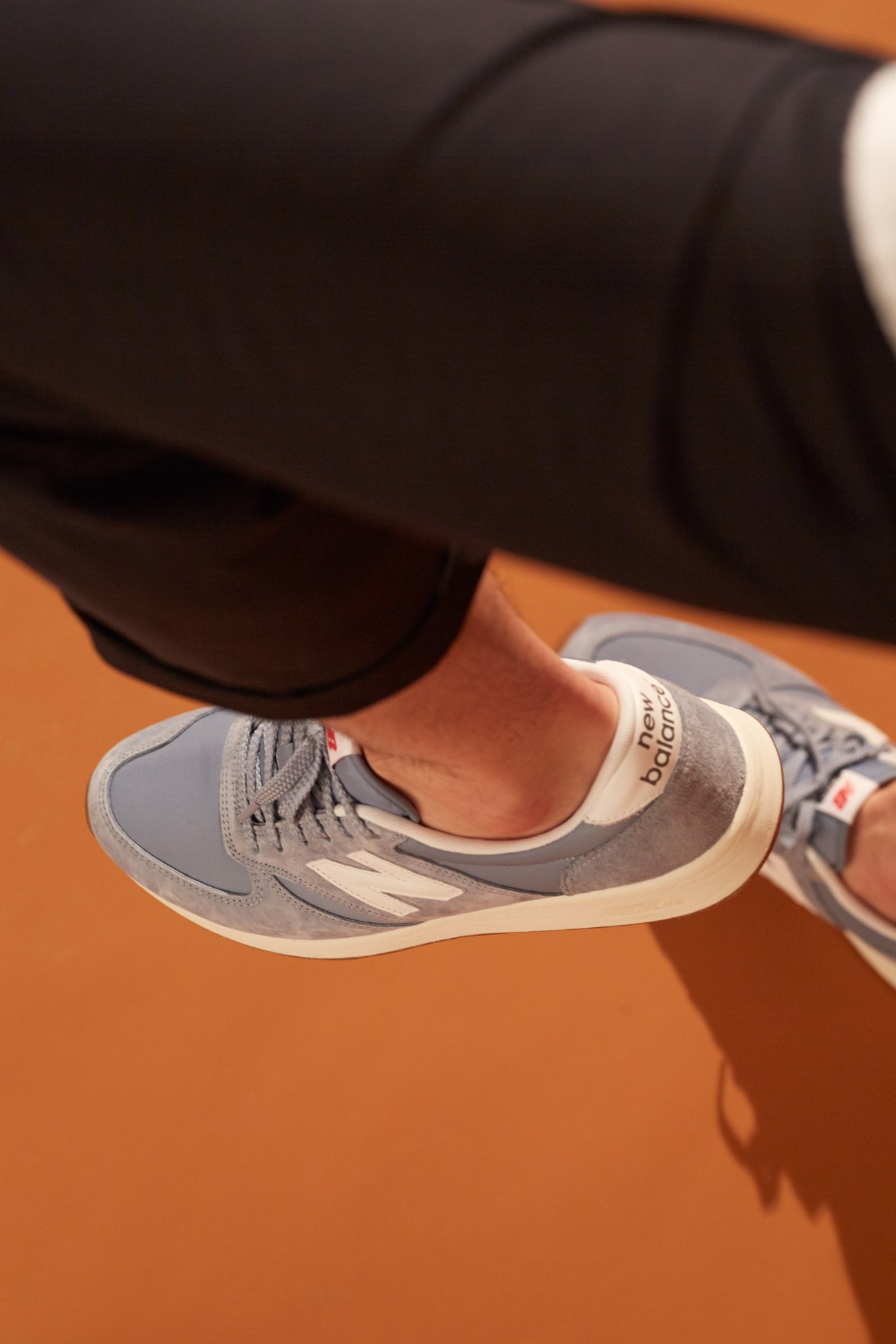 Grey and cream Heschung sneaker worn with black trousers on an orange background.