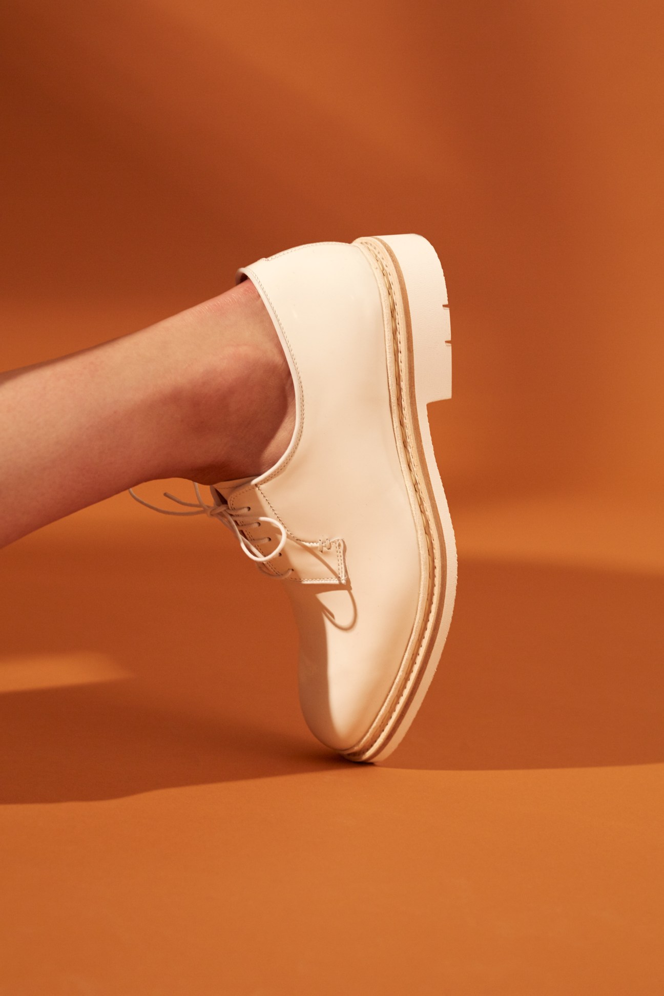 Cream-colored New Balance trainers with tan laces worn with bare leg against orange background.