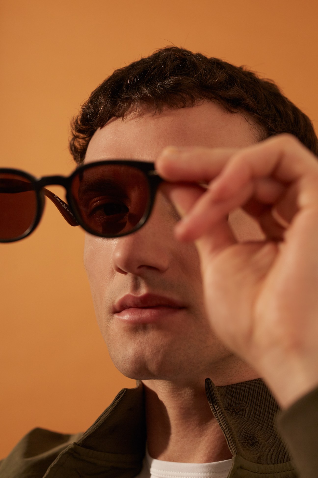 Man wearing Oliver Peoples pour Berluti sunglasses and Valstar jacket against orange background.