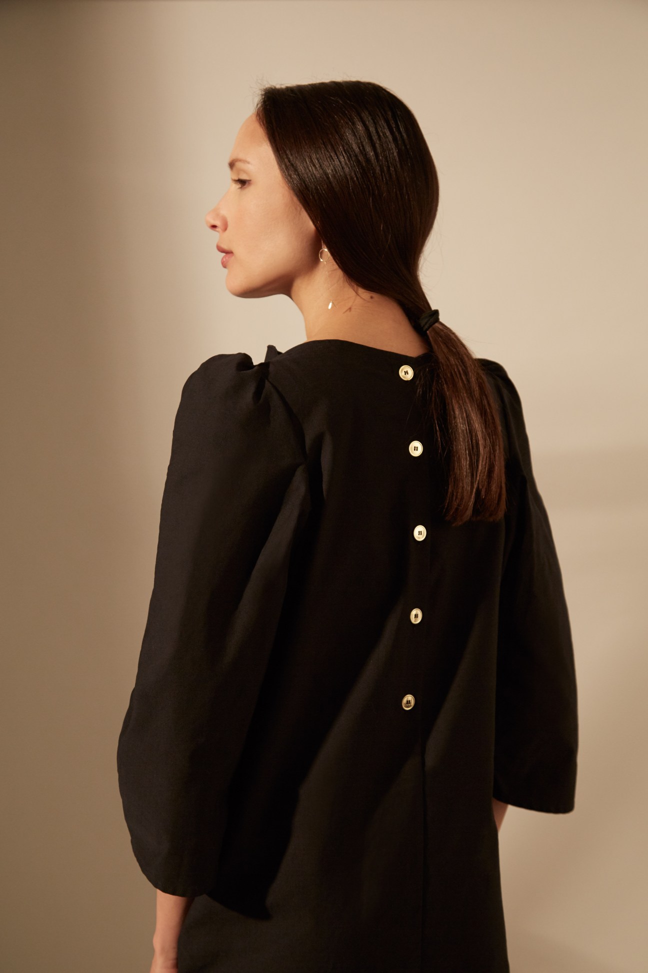 Woman wearing a black Atlantique Ascoli dress with puffed sleeves and gold buttons down the back, shown from behind.