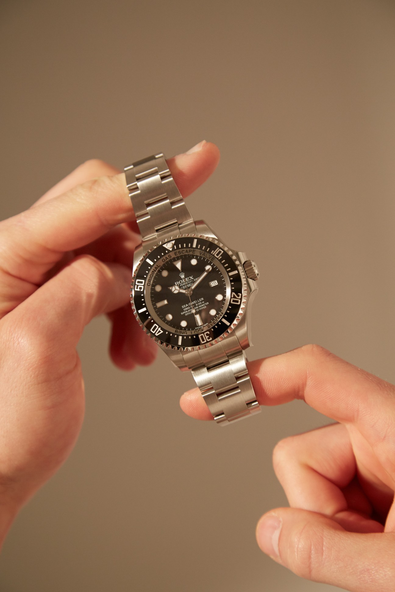 Hands holding a Rolex Sea-Dweller dive watch with black dial and steel bracelet against beige background.