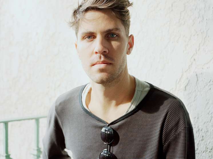 Portrait of Lo-Fang, a man with styled blonde and brown hair wearing a striped grey v-neck shirt with sunglasses hanging from the neckline.