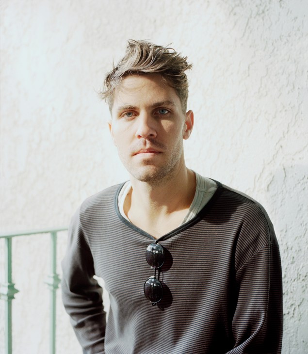 Portrait of Lo-Fang, a man with styled blonde and brown hair wearing a striped grey v-neck shirt with sunglasses hanging from the neckline.