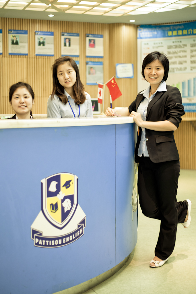 Three women staff members at Pattison English school reception desk with school logo visible.