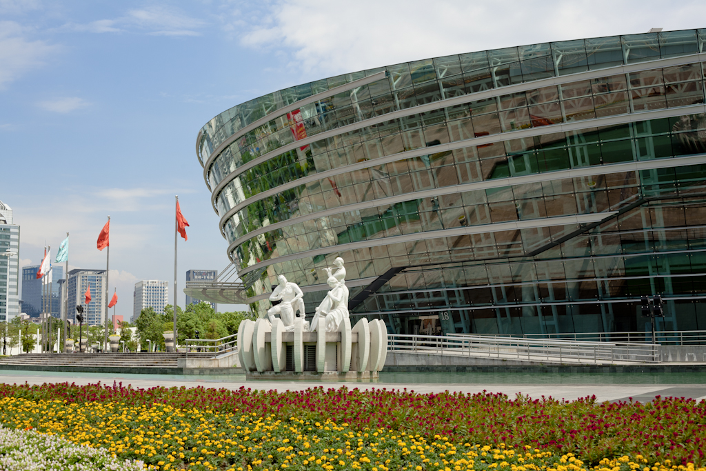 Modern circular building with glass facade beside sculptural figures in water and colorful flower beds in downtown Dongguan.