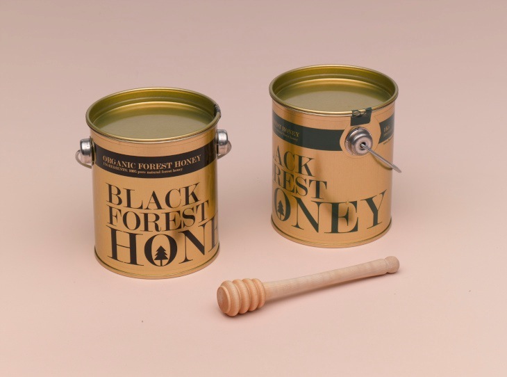 Two gold lacquered honey cans labeled 'Black Forest Honey' with a wooden honey dipper on a pink background.