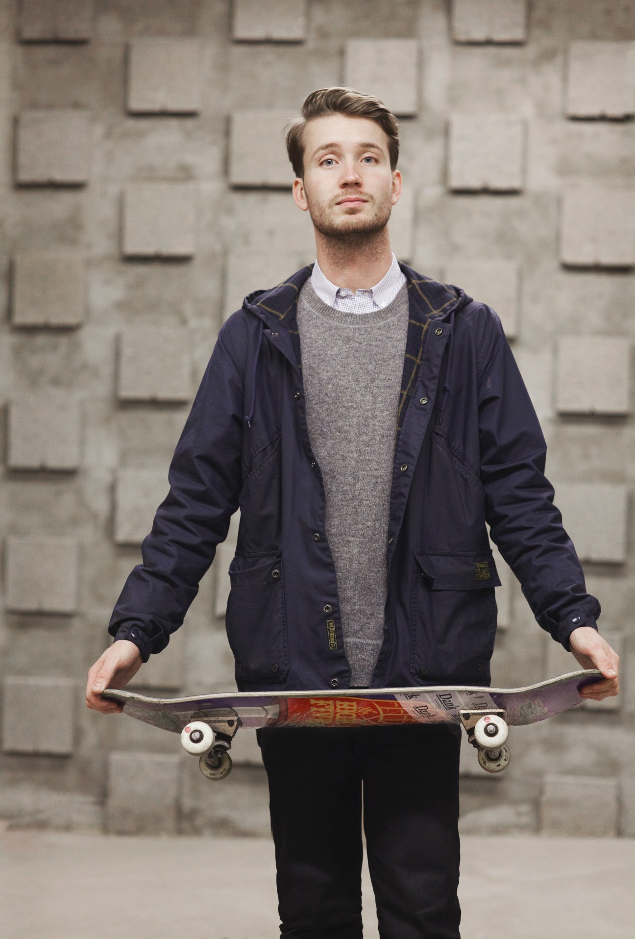Man holding skateboard against brick wall wearing navy jacket and gray sweater