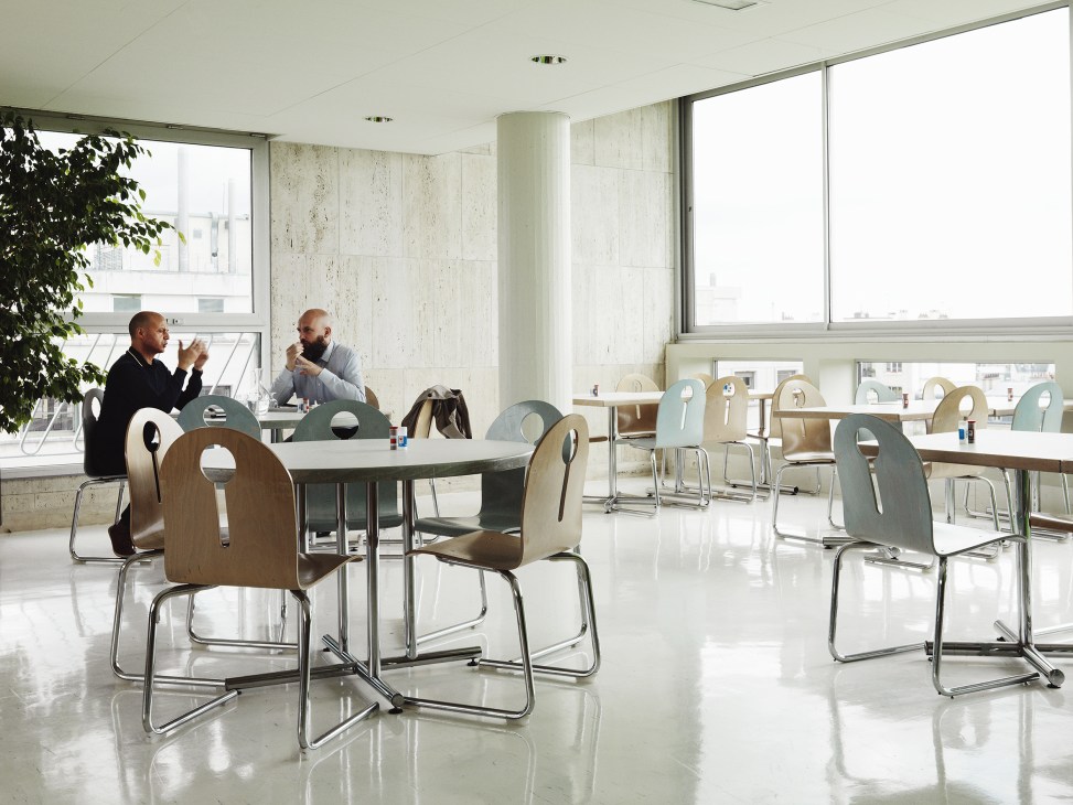 Two men converse at a table in a modernist cafeteria with distinctive wooden chairs and large windows overlooking Paris.