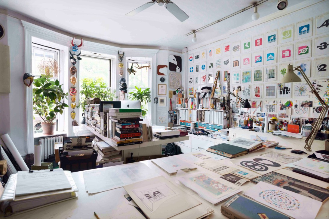 Lance Wyman's light-filled studio workspace with design sketches, samples, and architectural drawings spread across a table, surrounded by displayed logos and cultural artifacts on walls.