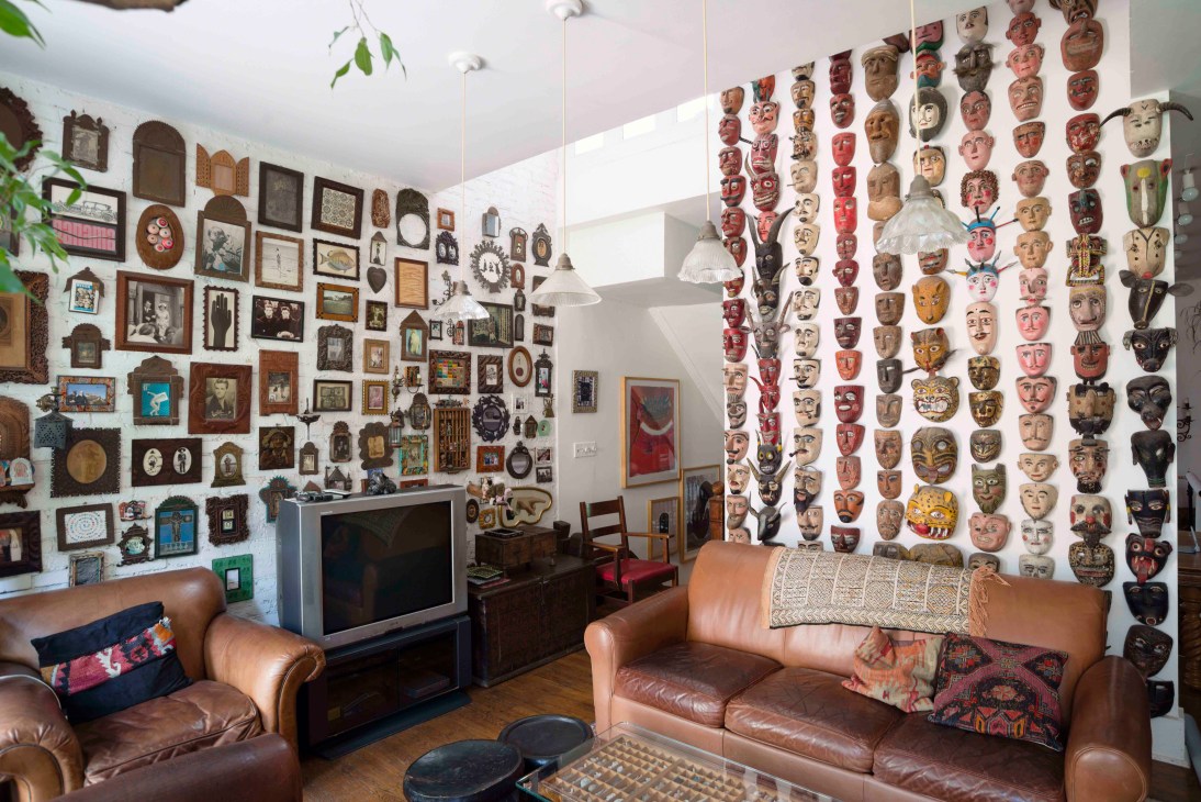 Lance Wyman's studio interior with walls covered in decorative masks, framed artwork, and cultural mementos alongside brown leather seating.
