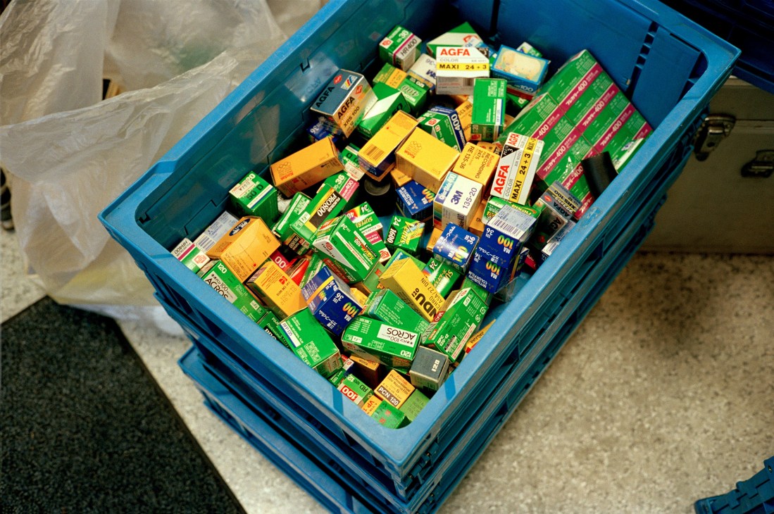 Blue crate filled with various colored film boxes including Agfa, Kodak, and other brands at a second-hand camera shop.