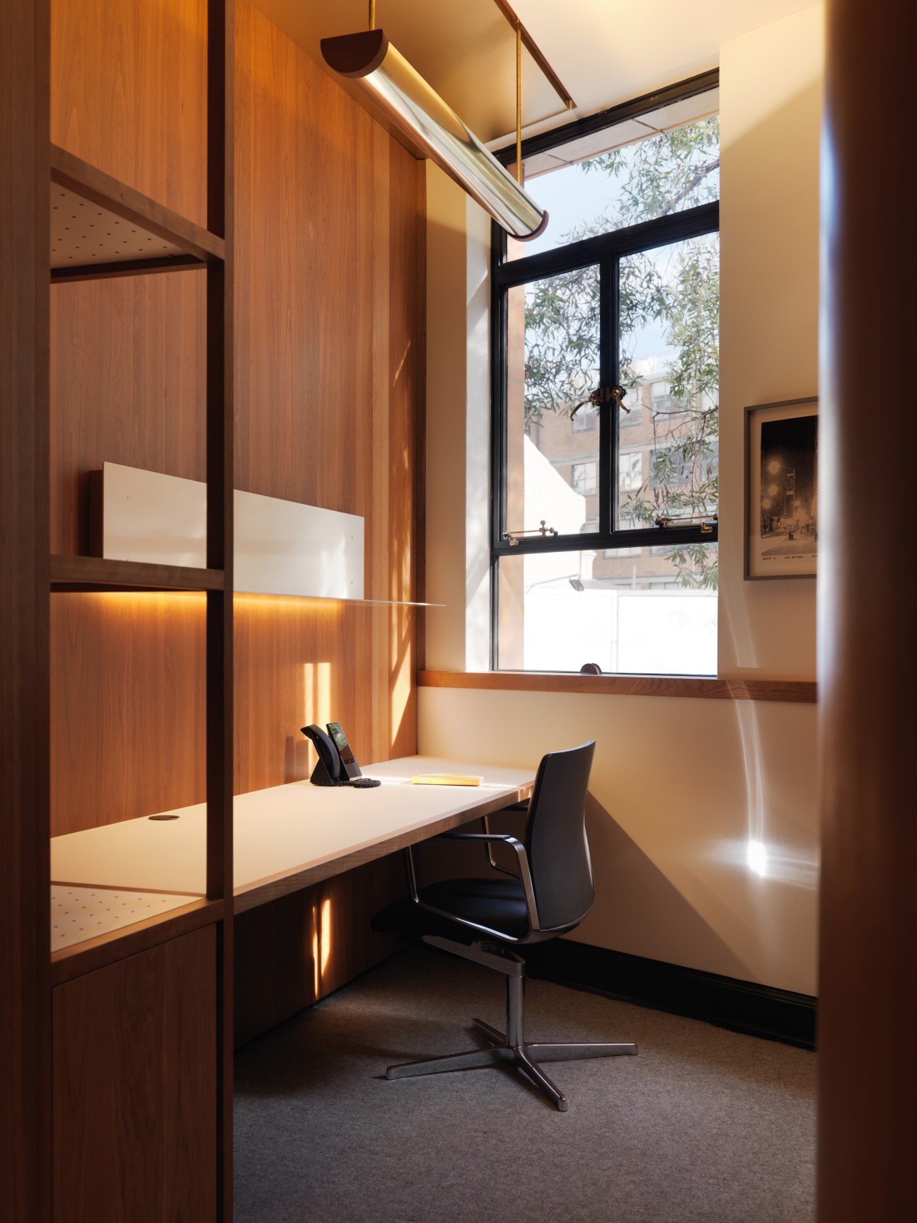 A minimalist office booth with dark cherry-wood joinery, a white desk, black chair, and large window overlooking a rooftop.