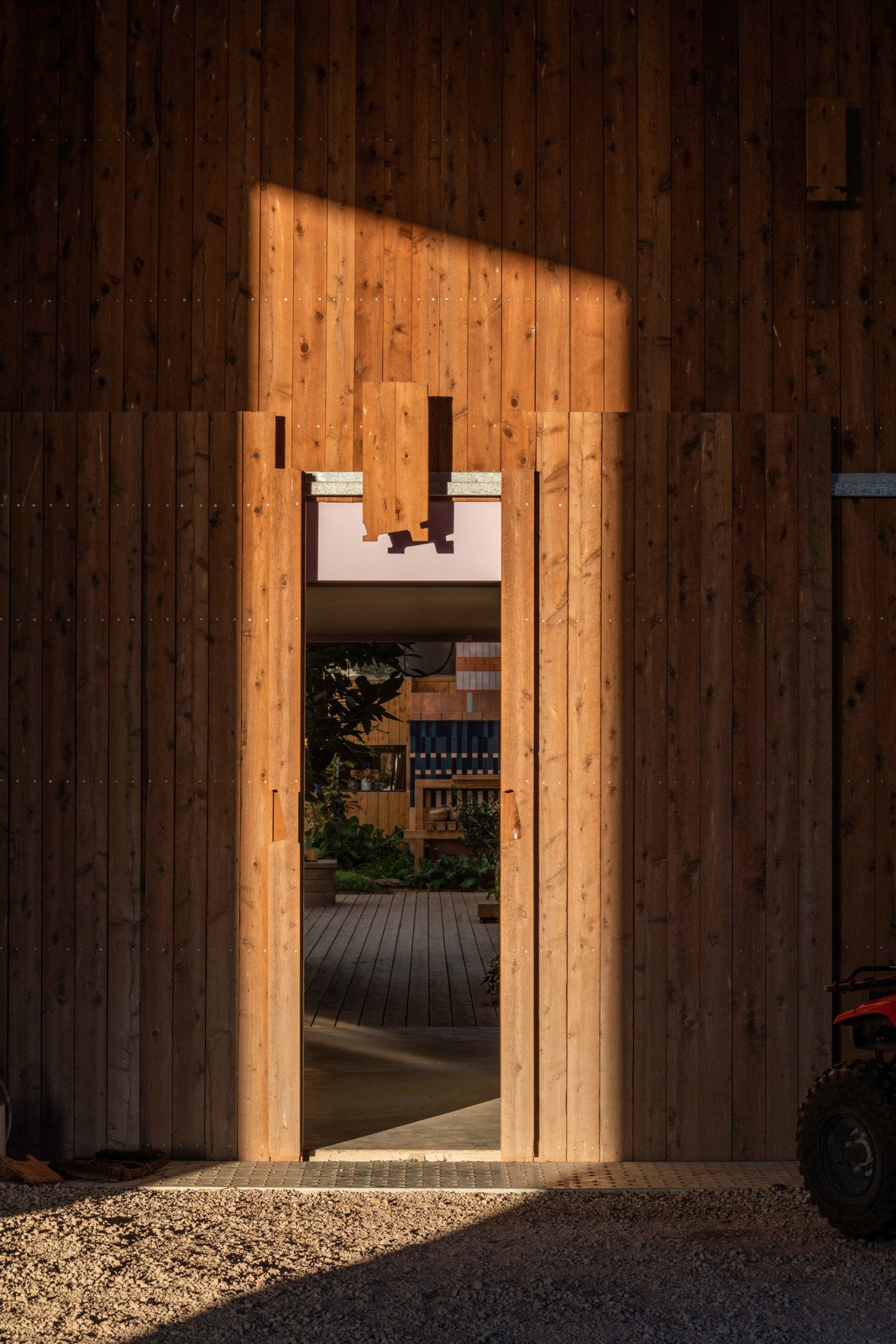 Sunlight illuminates a wooden doorway entrance revealing glimpses of an interior courtyard with timber decking and plants