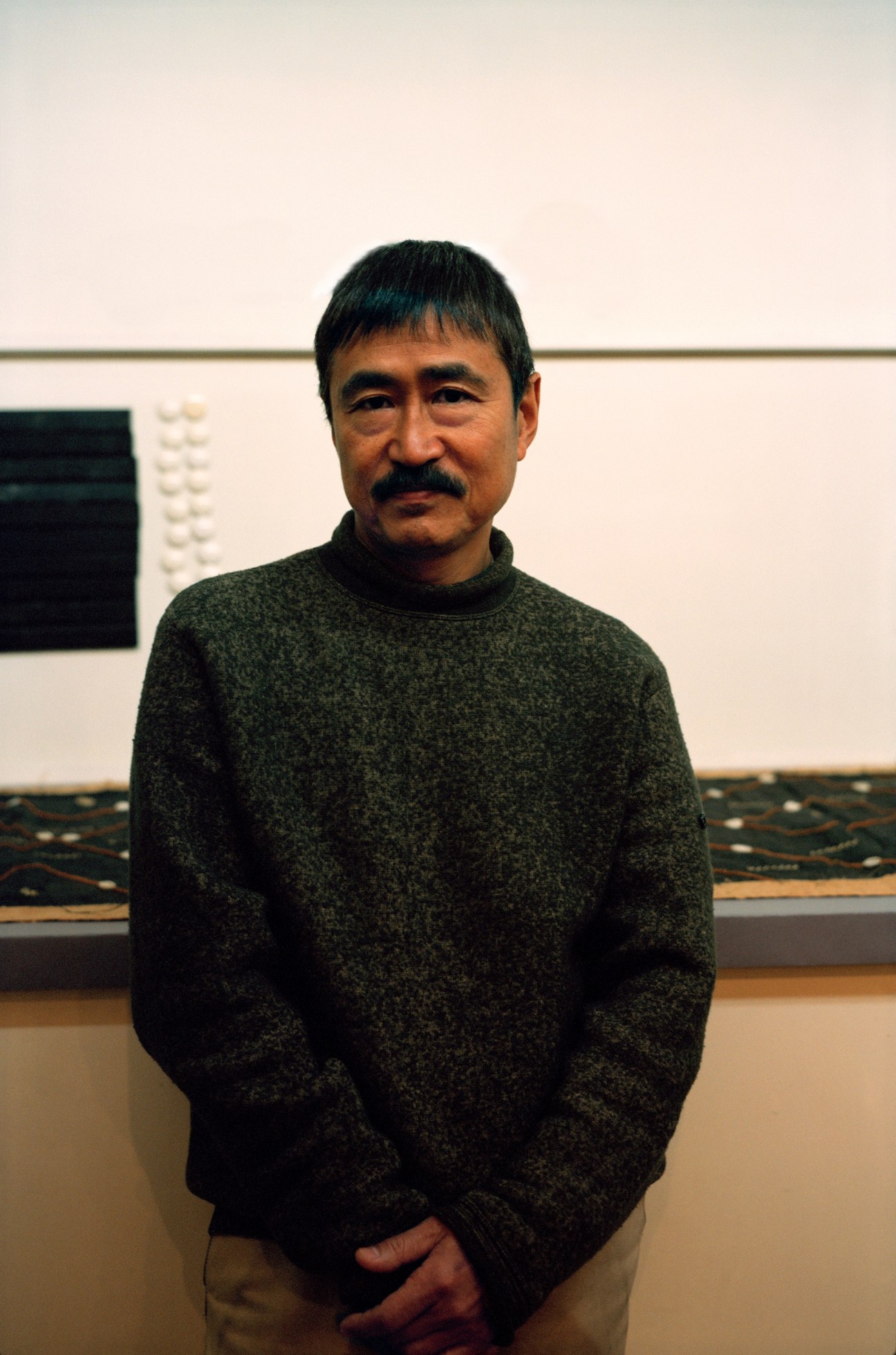 Photographer Taishi Hirokawa stands indoors wearing a green sweater, with framed photographs visible on the wall behind him.