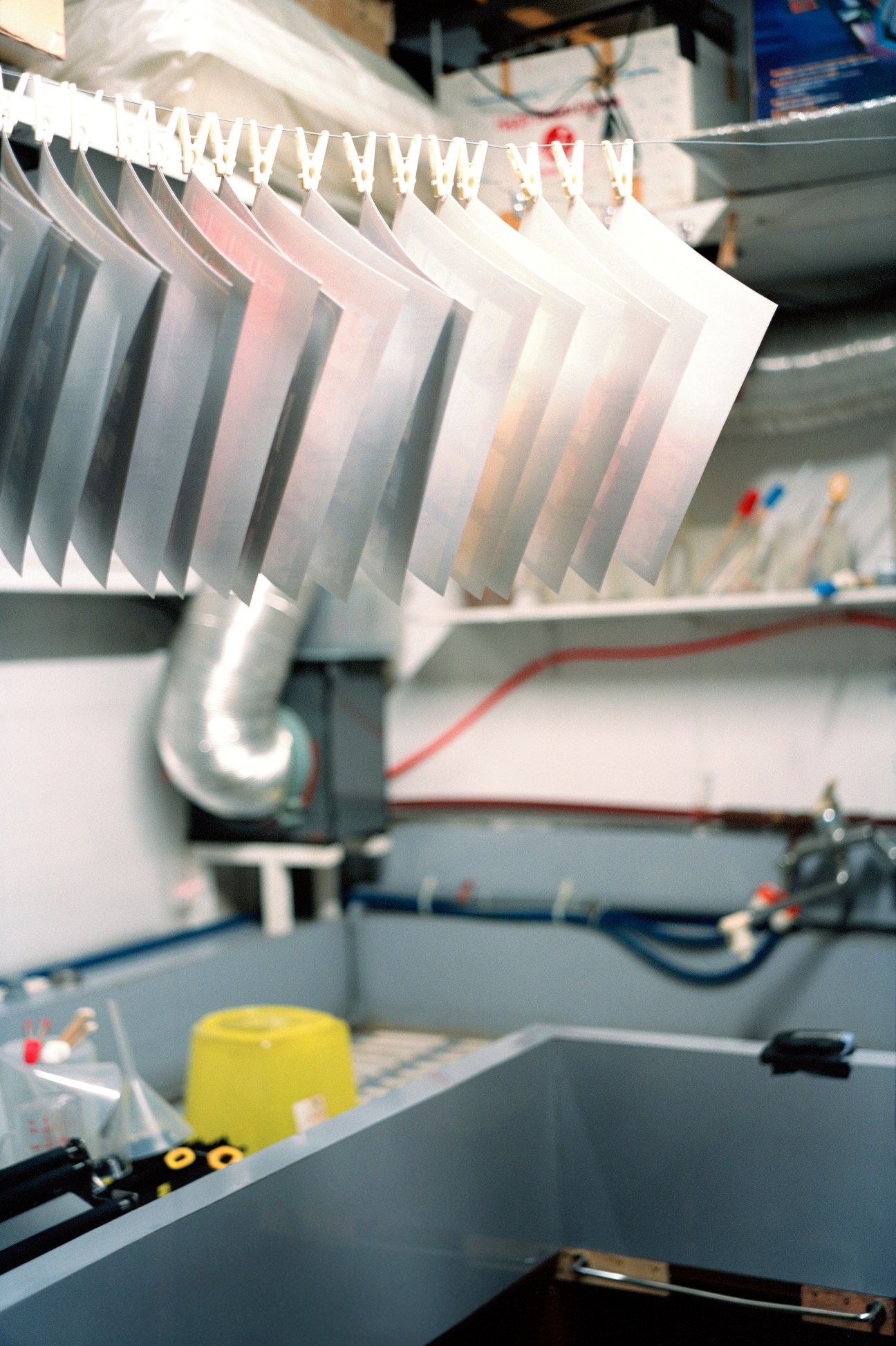 Photographic prints hang from a clothesline in a darkroom with chemical trays and equipment below.