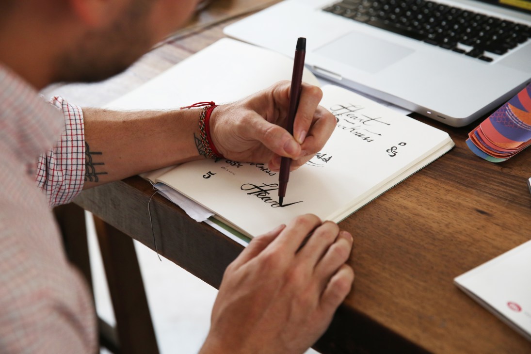 Person's hand writing in a notebook with a pencil at a desk with keyboard and tablet.