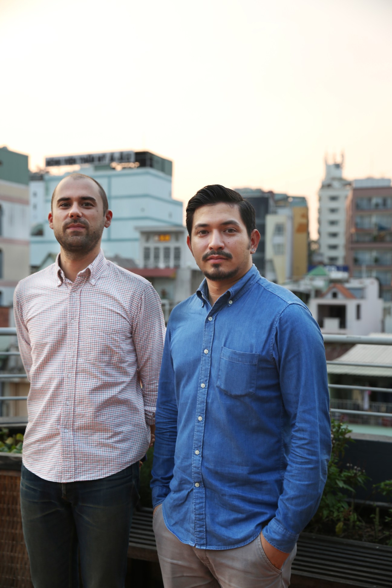 Two men stand on a rooftop with Ho Chi Minh City buildings in the background
