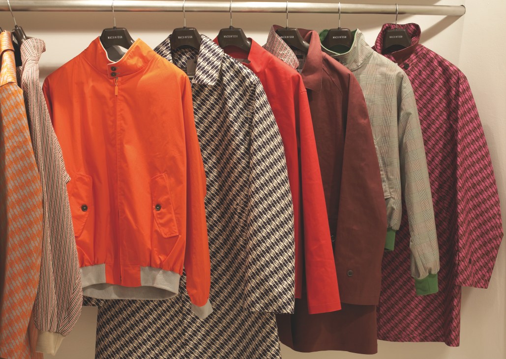 Collection of Mackintosh overcoats and jackets in vibrant colors including orange, red, black and patterned designs displayed on hangers.