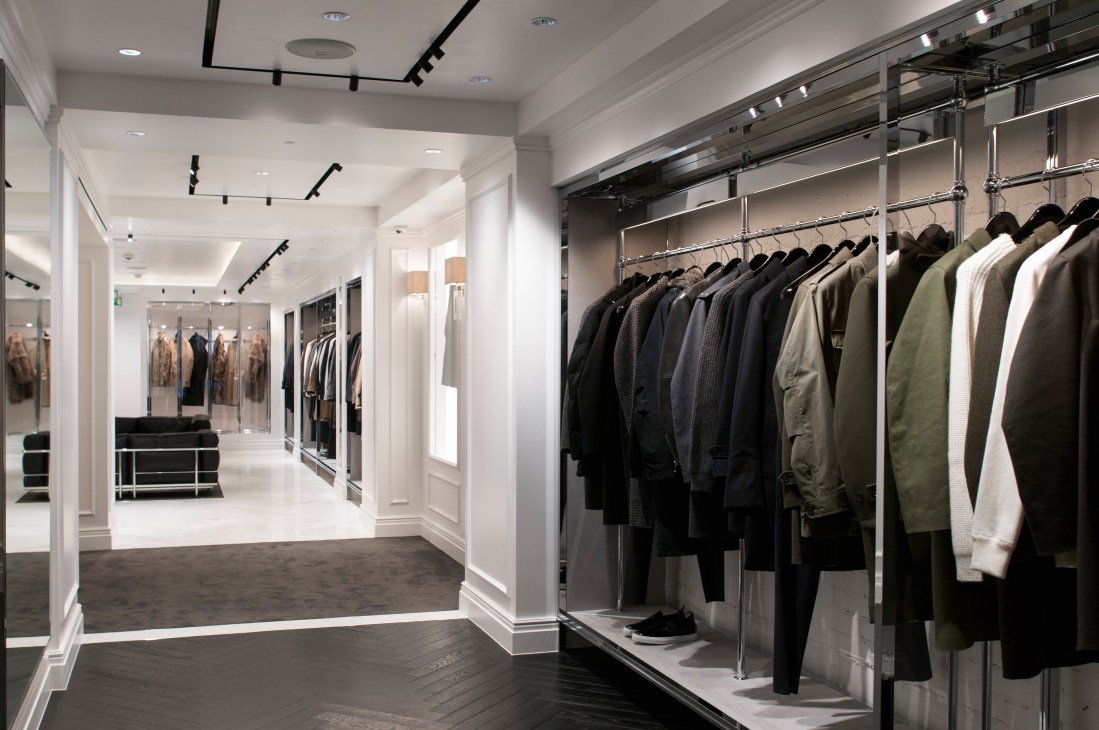 Interior of Mackintosh store on Conduit Street, London, showing minimalist white walls, chrome rails displaying coats and jackets, and polished dark flooring.
