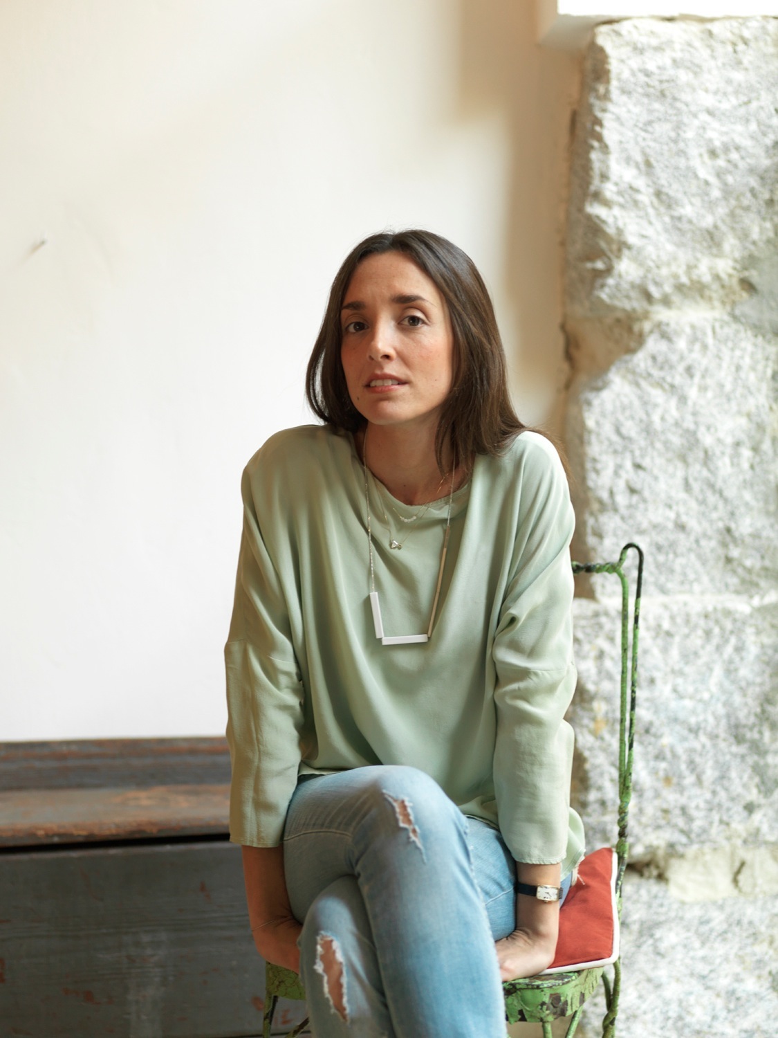 Woman in pale green long-sleeve shirt sitting on a colorful chair against a white brick wall.