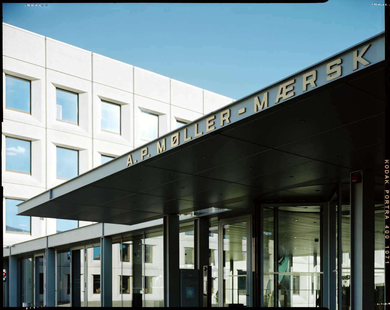 Maersk headquarters entrance with company name in gold lettering on dark canopy above glass doors and white marble facade.