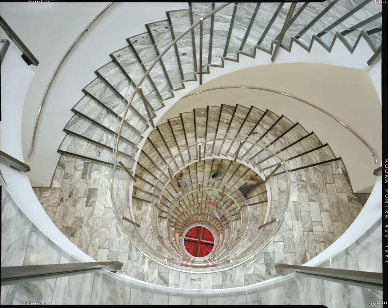Overhead view of a spiral staircase with metal railings descending multiple levels toward a red circular window at the center.