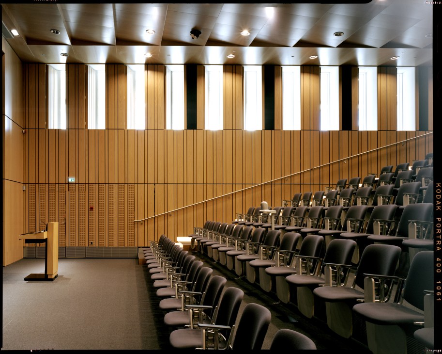 Oak-panelled auditorium with rows of black chairs, a wooden podium, and skylights in the ceiling.